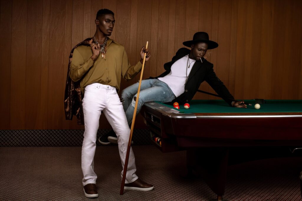Portrait of two handsome Black men posing by themselves inside a pool billiards hall playing pool both wearing casual but stylish wardrobe while smoking a joint and having a good time.