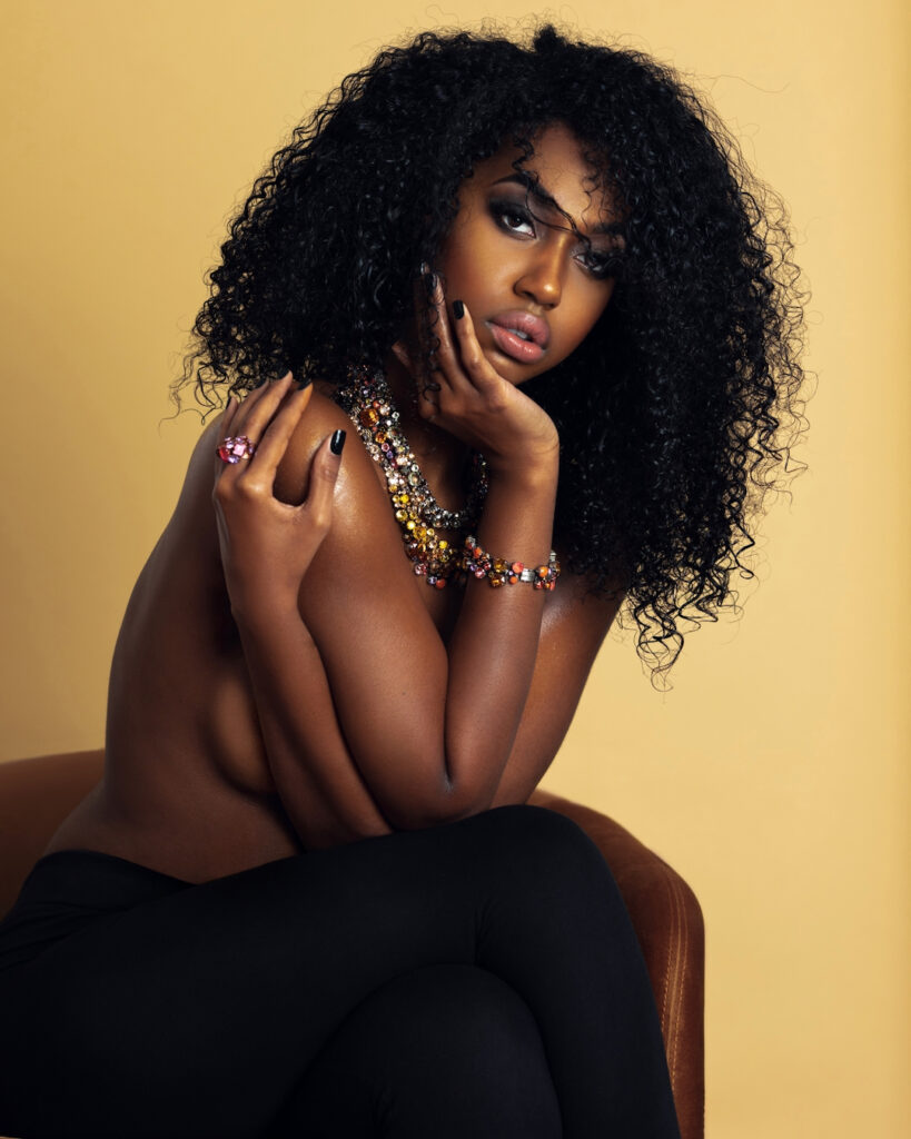 Portrait of a sensual Black woman with long curly hair and beautiful makeup sitting by herself on a golden velvet chair inside a studio with beige background wearing black pants with jewelry.