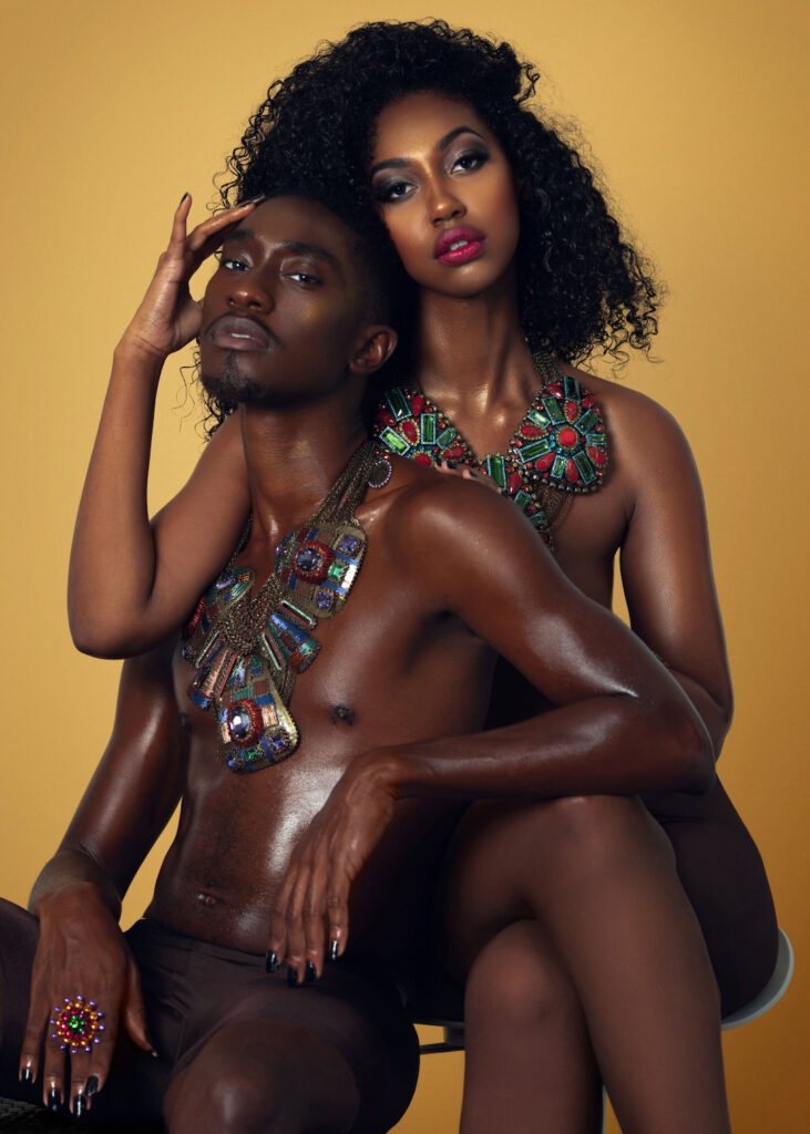 The Melanated Nation. Portrait of a beautiful Black couple with Afro hair and beautiful makeup sitting by themselves on chairs inside a studio with beige background wearing dark pants with large colorful jewelry.