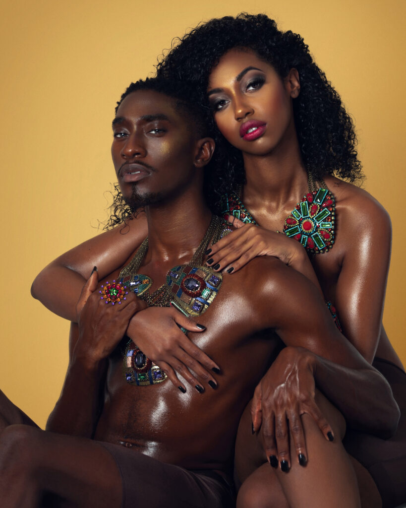 Portrait of a sensual Black couple with Afro hair and beautiful makeup sitting by themselves on chairs inside a studio with beige background wearing dark pants with large colorful jewelry.
