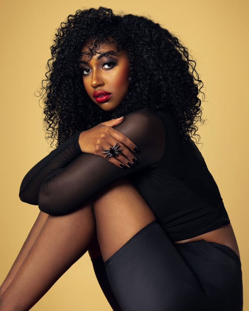 The Melanated Nation. Portrait of a beautiful Black woman with long curly hair and beautiful makeup sitting by herself on a chair inside a studio with beige background wearing black pants with jewelry.