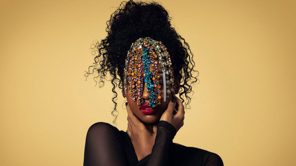 Closeup portrait of a Black woman with long curly hair and beautiful makeup posing by herself inside a studio with beige background wearing a black top with jewelry covering her face.