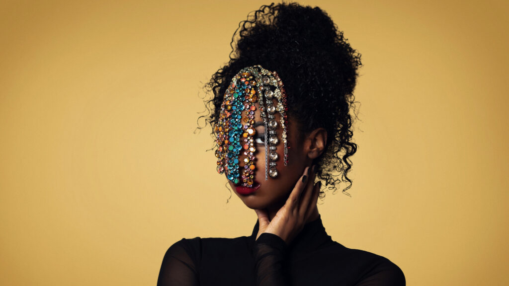 Closeup portrait of a confident Black woman with long curly hair and beautiful makeup posing by herself inside a studio with beige background wearing a black top with jewelry covering her face.