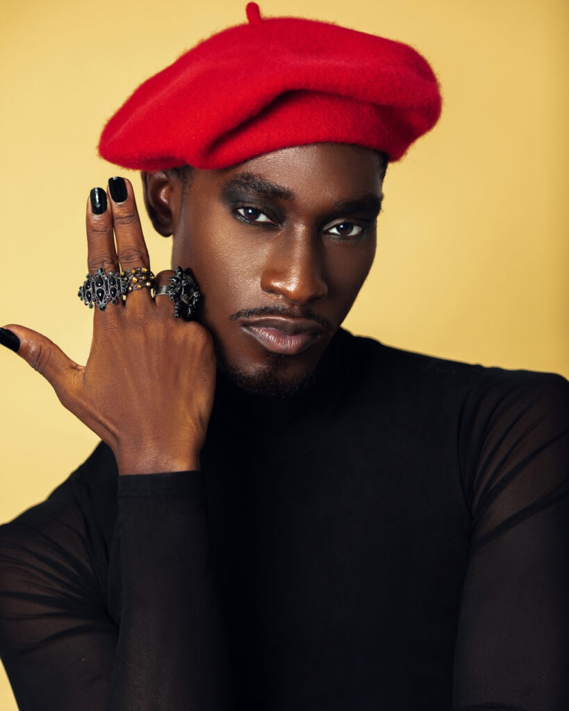 The Melanated Nation. Closeup portrait of a confident Black man with light makeup posing by himself inside a studio with beige background wearing a black top with jewelry and a red French barret.