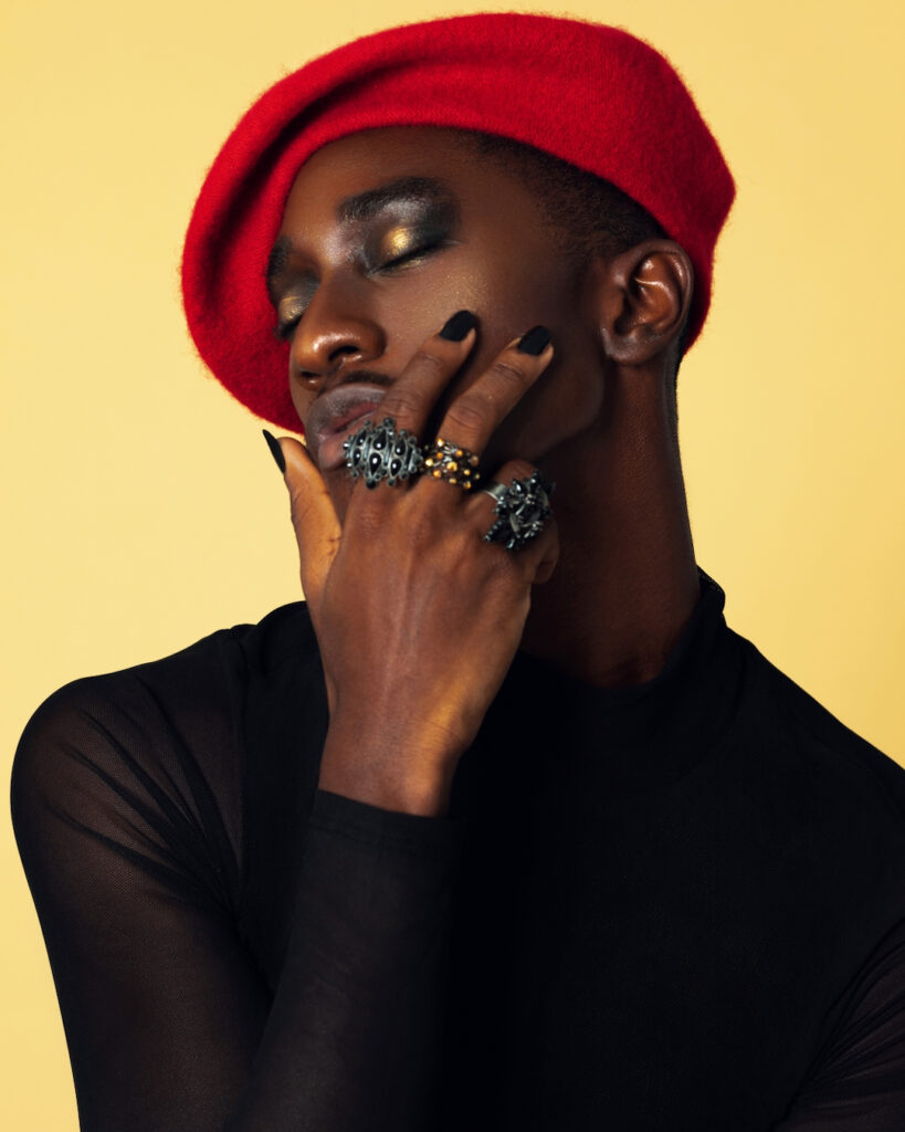 Closeup portrait of a serene Black man with light makeup posing by himself inside a studio with beige background wearing a black top with jewelry and a red French barret.