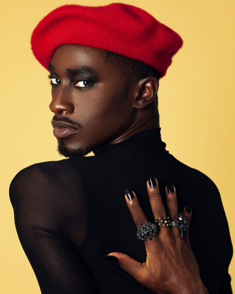 Closeup portrait of a handsome Black man with light makeup posing by himself inside a studio with beige background wearing a black top with jewelry and a red French barret.