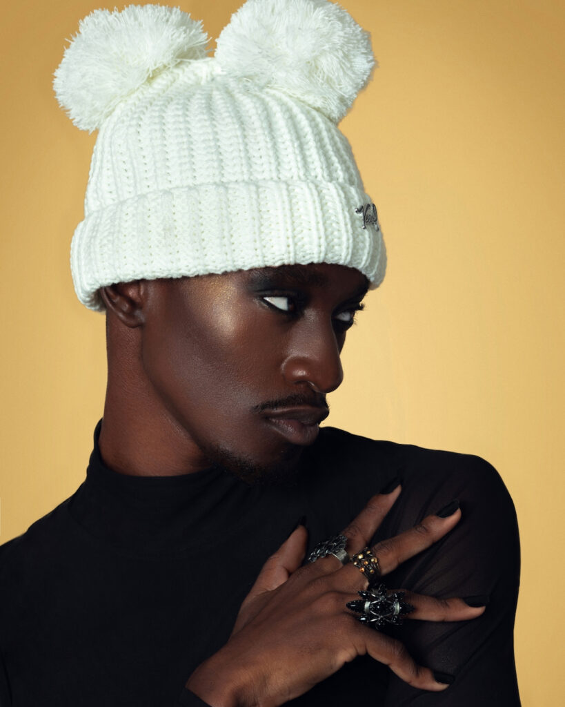 Closeup portrait of a serene Black man with light makeup posing by himself inside a studio with beige background wearing a black top with jewelry and a white ski hat.