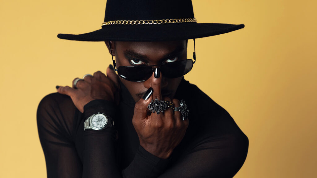 The Melanated Nation. Closeup portrait of a confident Black man with light makeup posing by himself inside a studio with beige background wearing a black top with jewelry, a black fedora hat and sunglasses.
