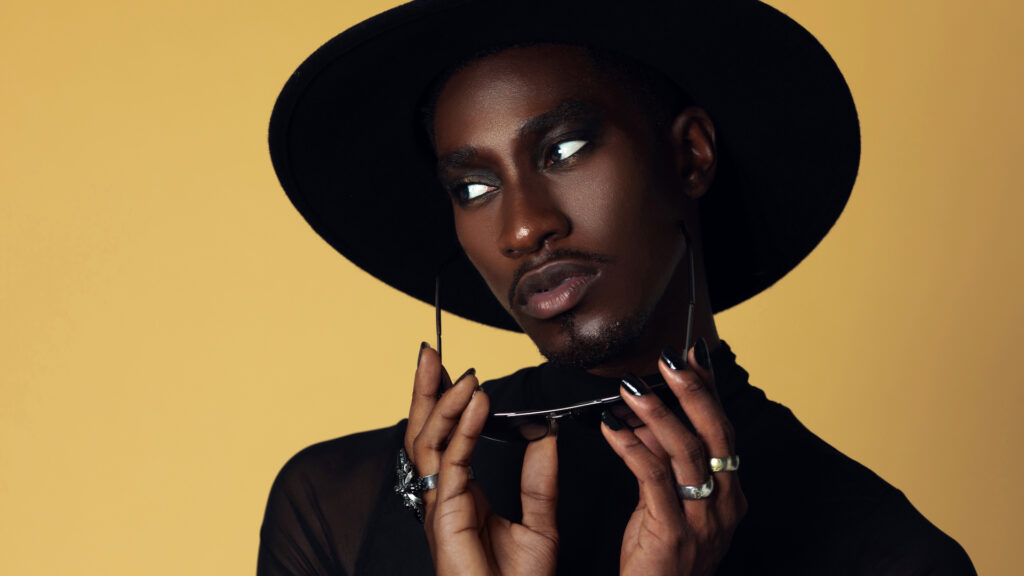 The Melanated Nation. Closeup portrait of a serene Black man with light makeup posing by himself inside a studio with beige background wearing a black top with jewelry, a black fedora hat and sunglasses.