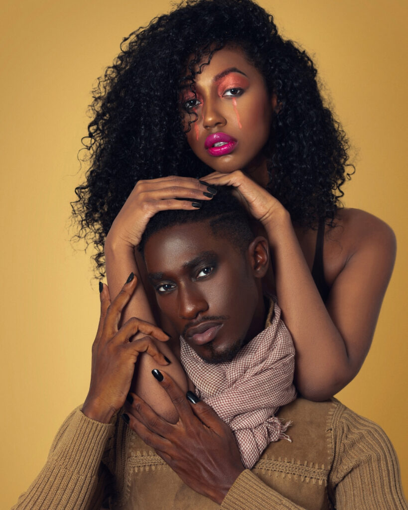 The Melanated Nation. Portrait of a beautiful Black couple with Afro hair and beautiful makeup sitting by themselves on chairs inside a studio with beige background wearing a beige sweater with a scarf.