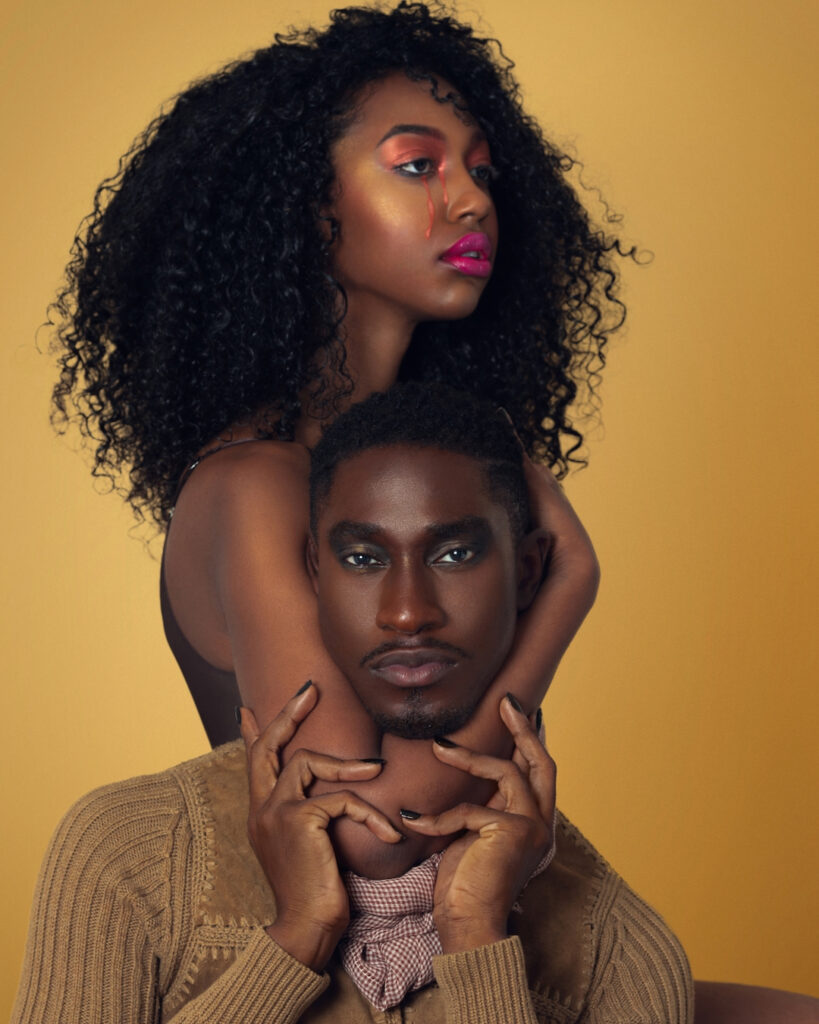 Portrait of a serene Black couple with Afro hair and beautiful makeup sitting by themselves on chairs inside a studio with beige background wearing a beige sweater with a scarf.
