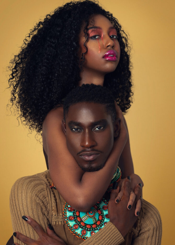 Portrait of a sensual Black couple with Afro hair and beautiful makeup sitting by themselves on chairs inside a studio with beige background wearing a beige sweater with jewelry.