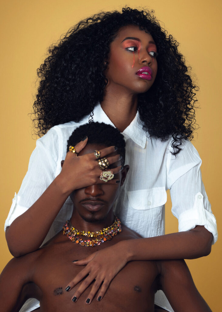 The Melanated Nation. Portrait of a gorgeous Black couple with Afro hair and beautiful makeup sitting by themselves on chairs inside a studio with beige background wearing a white shirt with jewelry.