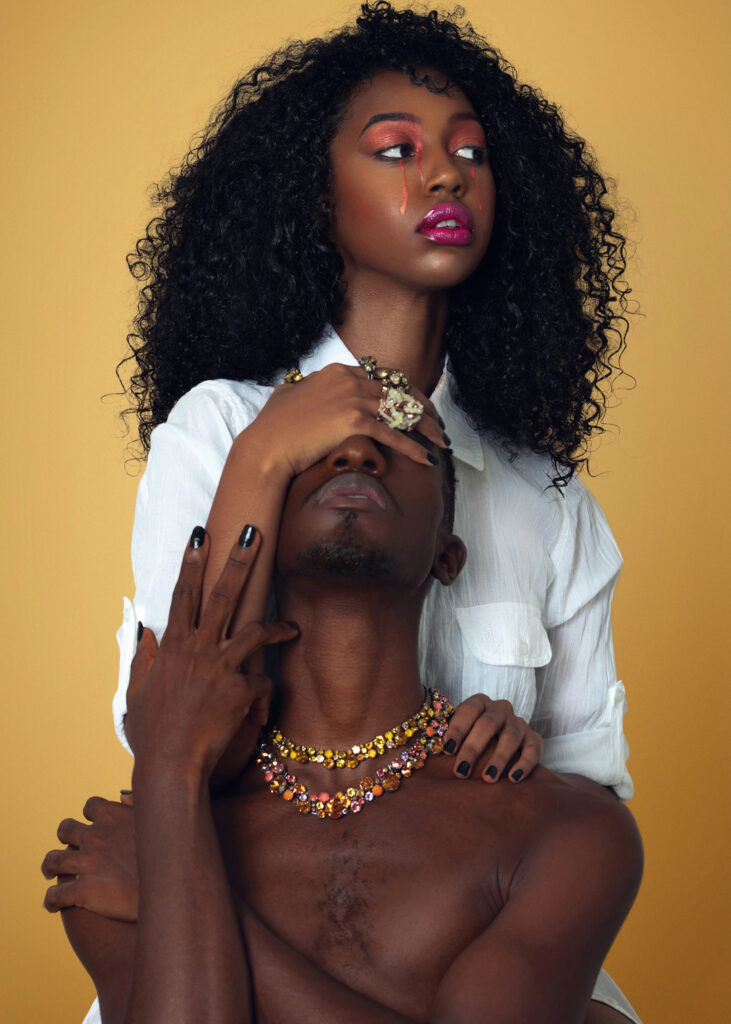 Portrait of a scared Black couple with Afro hair and beautiful makeup sitting by themselves on chairs inside a studio with beige background wearing a white shirt with jewelry.