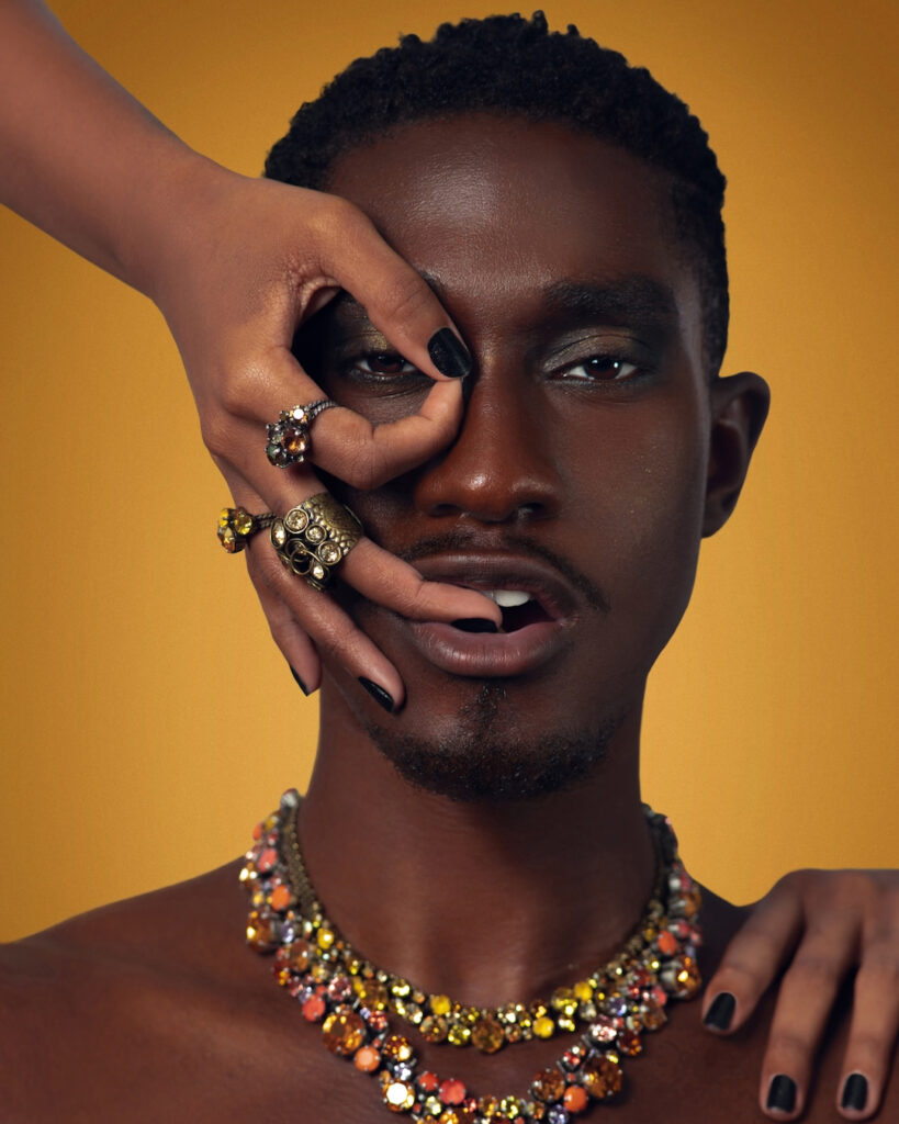 Closeup portrait of a confident Black man with short Afro hair and beautiful makeup posing by himself inside a studio with beige background wearing jewelry and a female hand covering his head.
