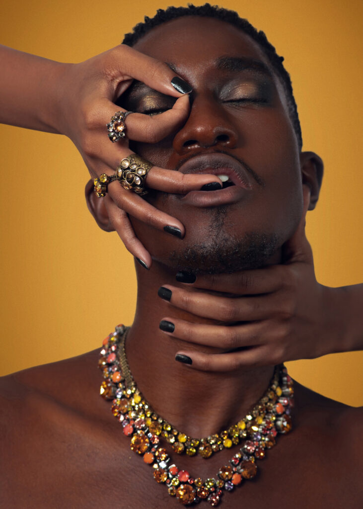 Closeup portrait of a serene Black man with short Afro hair and beautiful makeup posing by himself inside a studio with beige background wearing jewelry and a female hand covering his head.