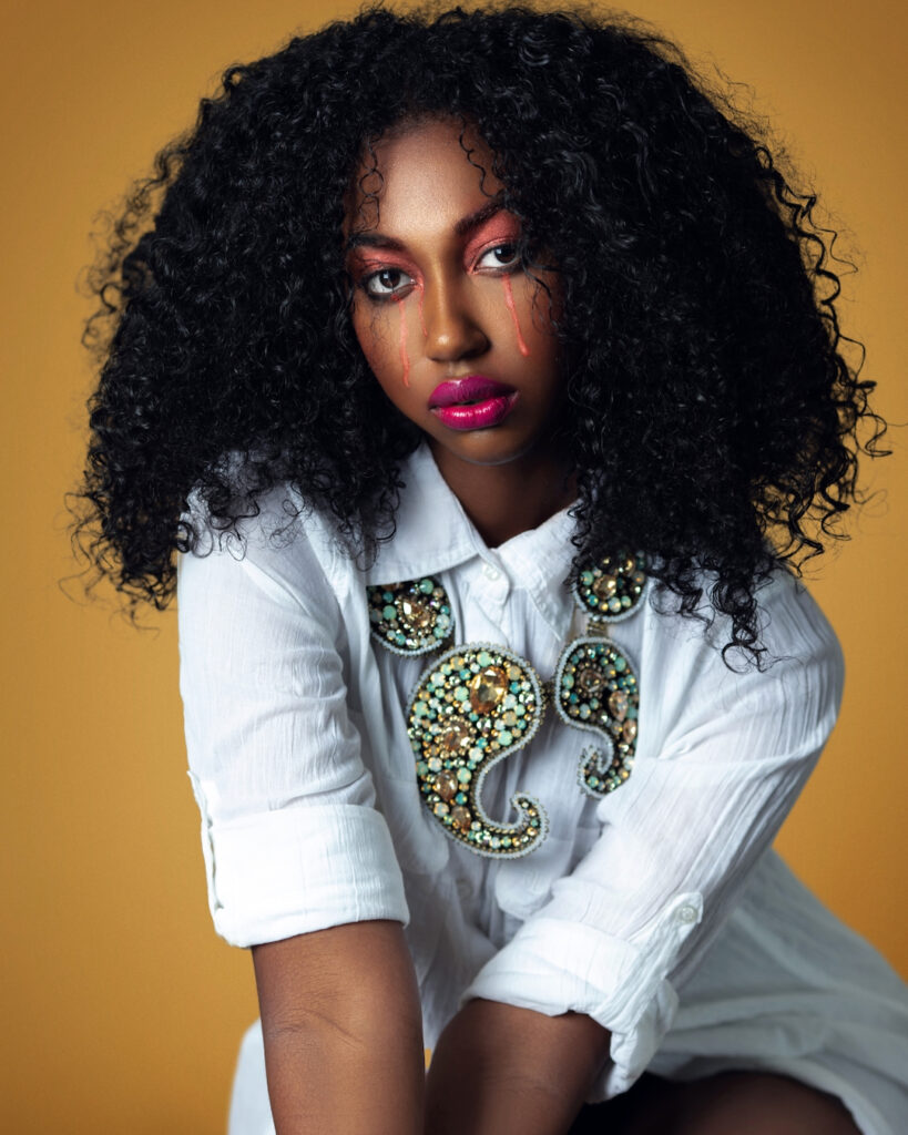 The Melanated Nation. Closeup portrait of a serene Black woman with long curly hair and beautiful makeup posing by herself inside a studio with beige background wearing a white shirt with jewelry.