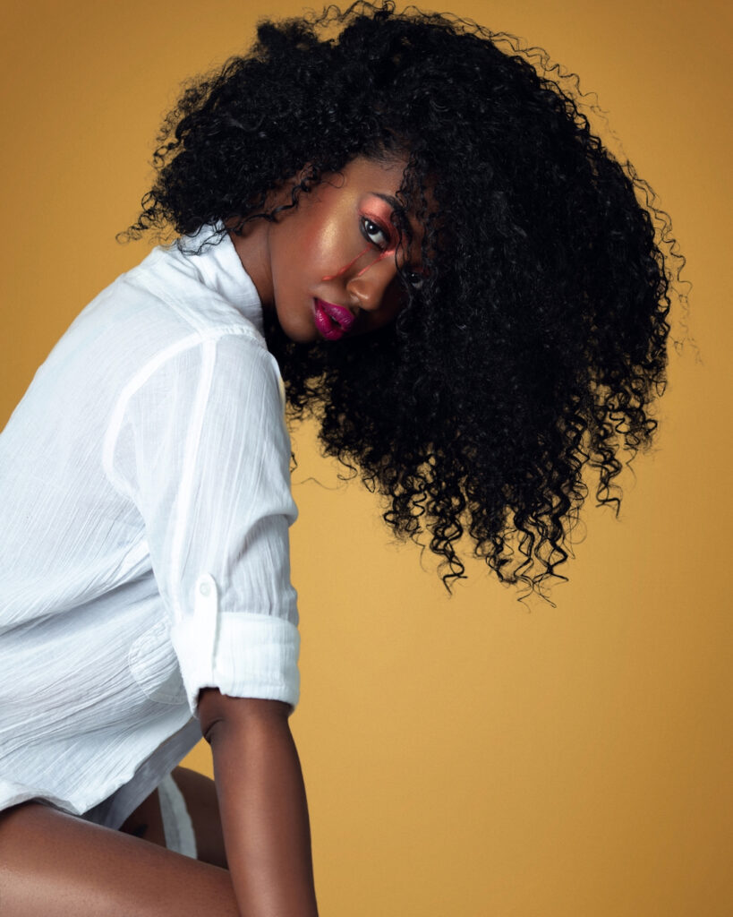 Closeup portrait of a sensual Black woman with long curly hair and beautiful makeup posing by herself inside a studio with beige background wearing a white shirt with jewelry.