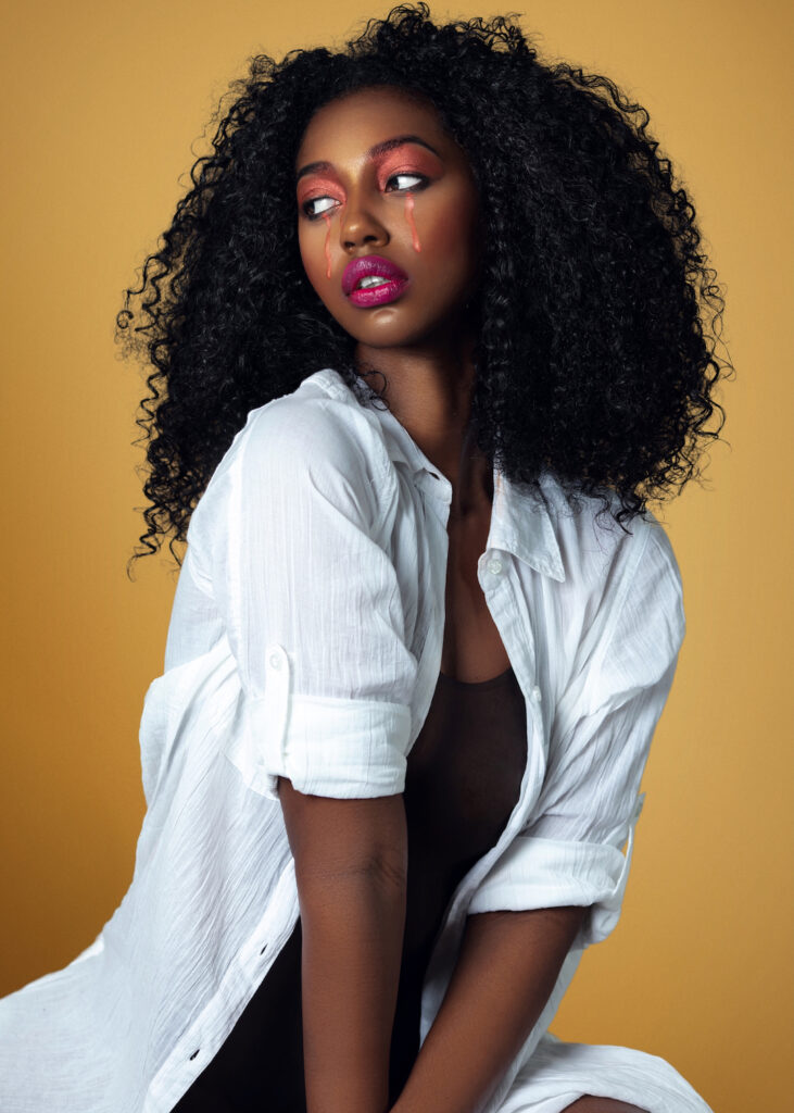 Closeup portrait of a serene Black woman with long curly hair and beautiful makeup posing by herself inside a studio with beige background wearing a white shirt with jewelry.