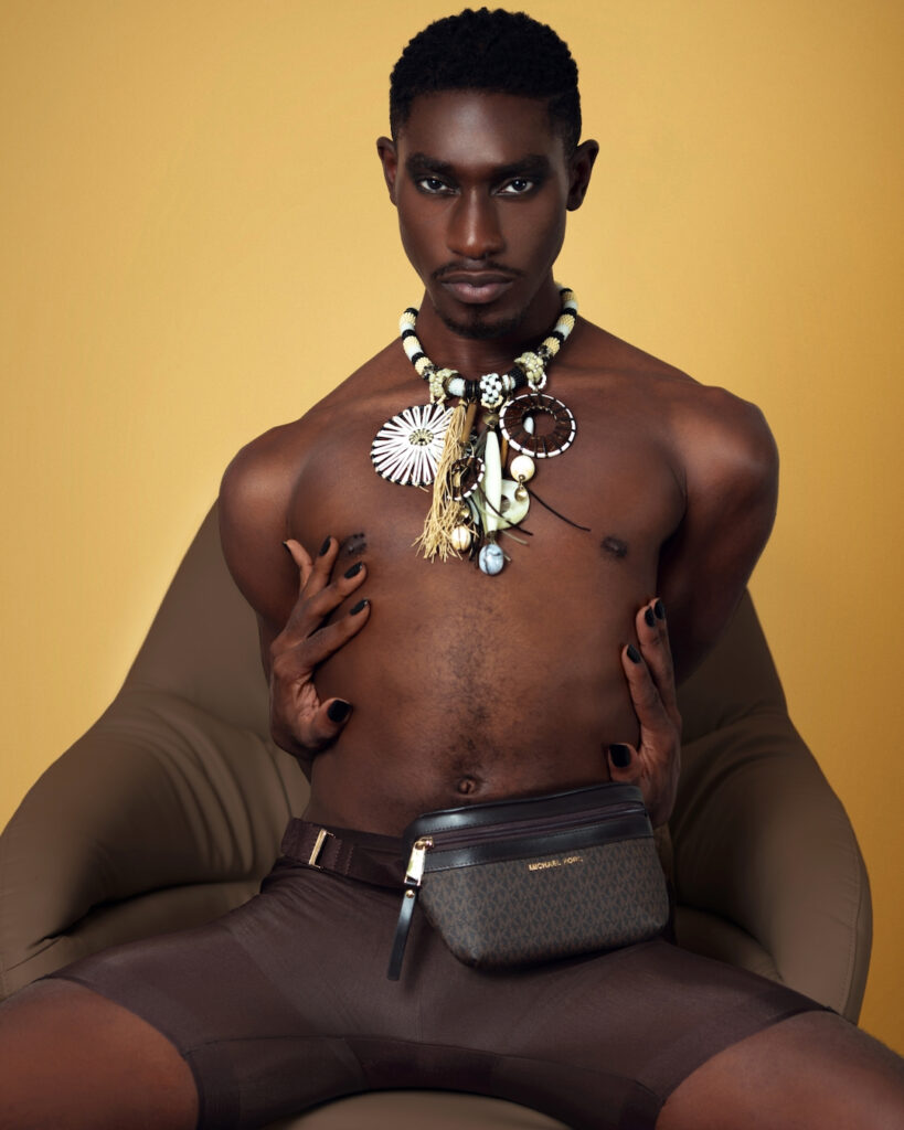 Portrait of a sensual Black man with short Afro hair and beautiful makeup sitting by himself on a grey leather lounge chair inside a studio with beige background wearing a shirt, shorts and jewelry.