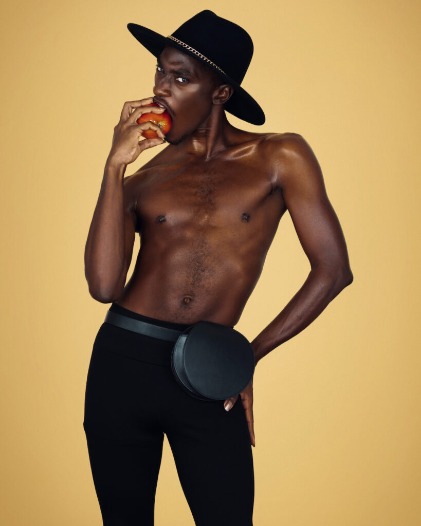 The Melanated Nation. Portrait of a strong Black man with a black hat and light makeup biting into an apple posing by himself inside a studio with beige background wearing black pants with a purse on a belt.