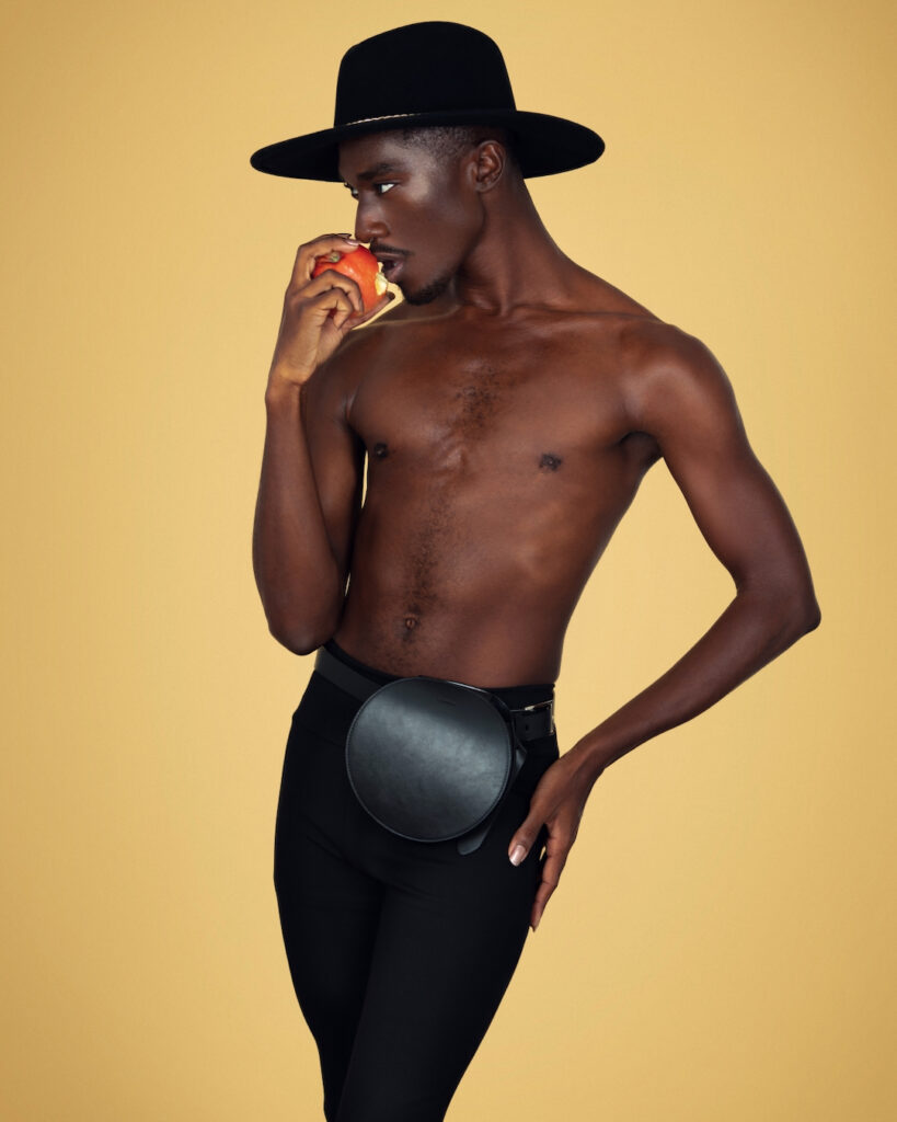 Portrait of a serene Black man with a black hat and light makeup eating an apple posing by himself inside a studio with beige background wearing black pants with a purse on a belt.