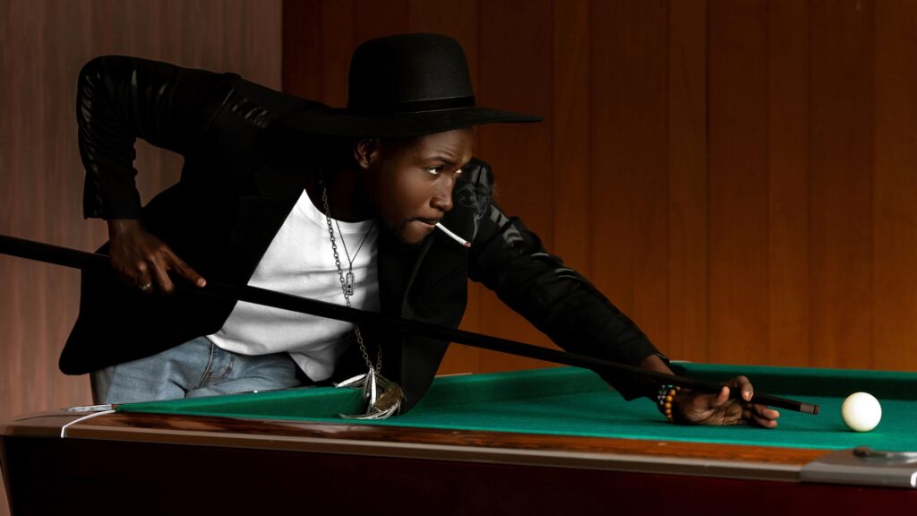 billiards wearing a black leather jacket, white T-shirt with with blue denim jeans and a long necklace.