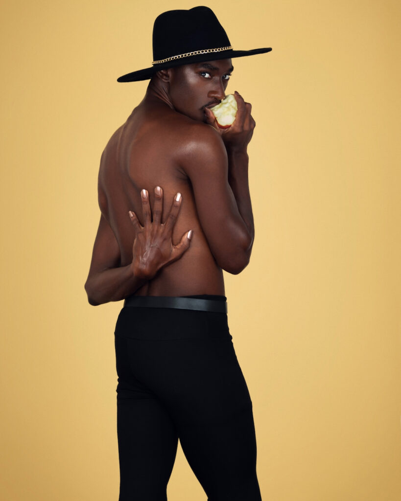 Portrait of a beautiful Black man with a black hat and light makeup eating an apple posing with one hand on his back inside a studio with beige background wearing black pants with a purse on a belt.