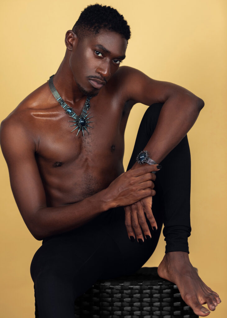 Portrait of a confident Black man with faded Afro hair and light makeup sitting by himself on a cube inside a studio with beige background wearing black pants with jewelry.