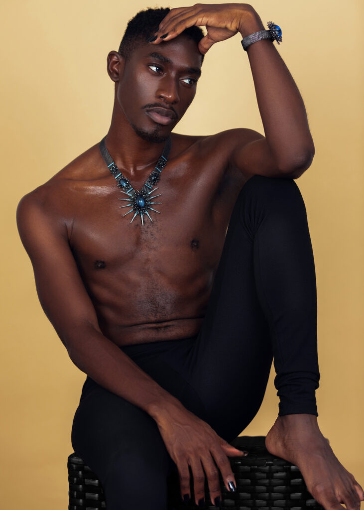 Portrait of a serene Black man with faded Afro hair and light makeup sitting by himself on a cube inside a studio with beige background wearing black pants with jewelry.