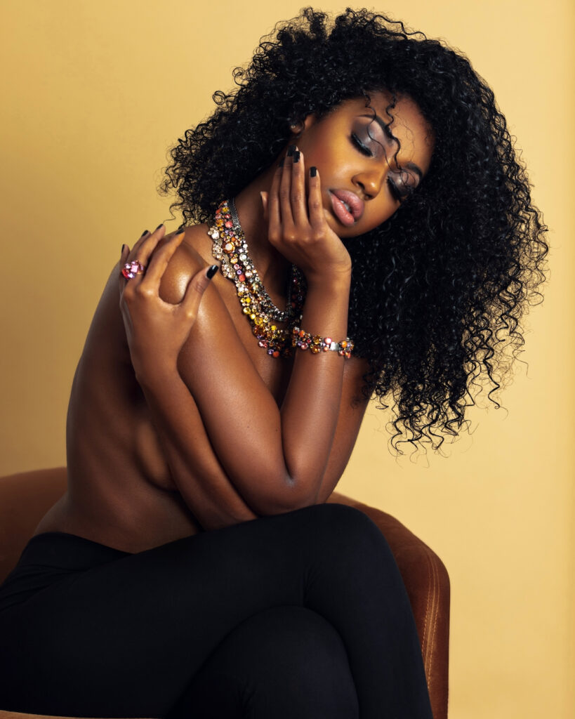Portrait of a serene Black woman with long curly hair and beautiful makeup sitting by herself on a golden velvet chair inside a studio with beige background wearing black pants with jewelry.