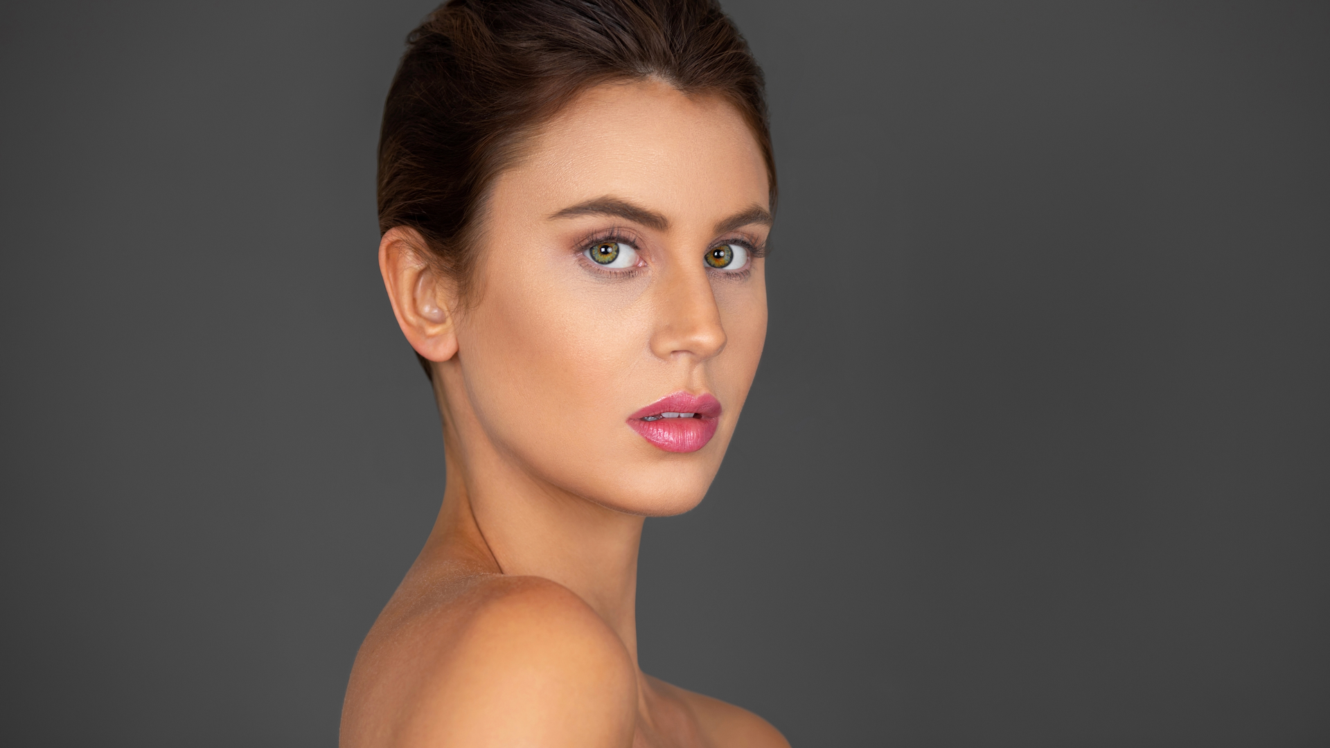 Portrait headshot of a vulnerable young white woman with wavy hair in a bun and beautiful makeup and lipstick posing by herself inside a studio with a grey background.