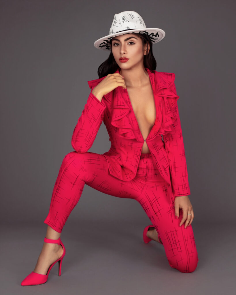 Portrait of a beautiful young white woman with wavy black hair and beautiful makeup posing by herself inside a studio with a grey background wearing a red business suit with high heels and a white hat.