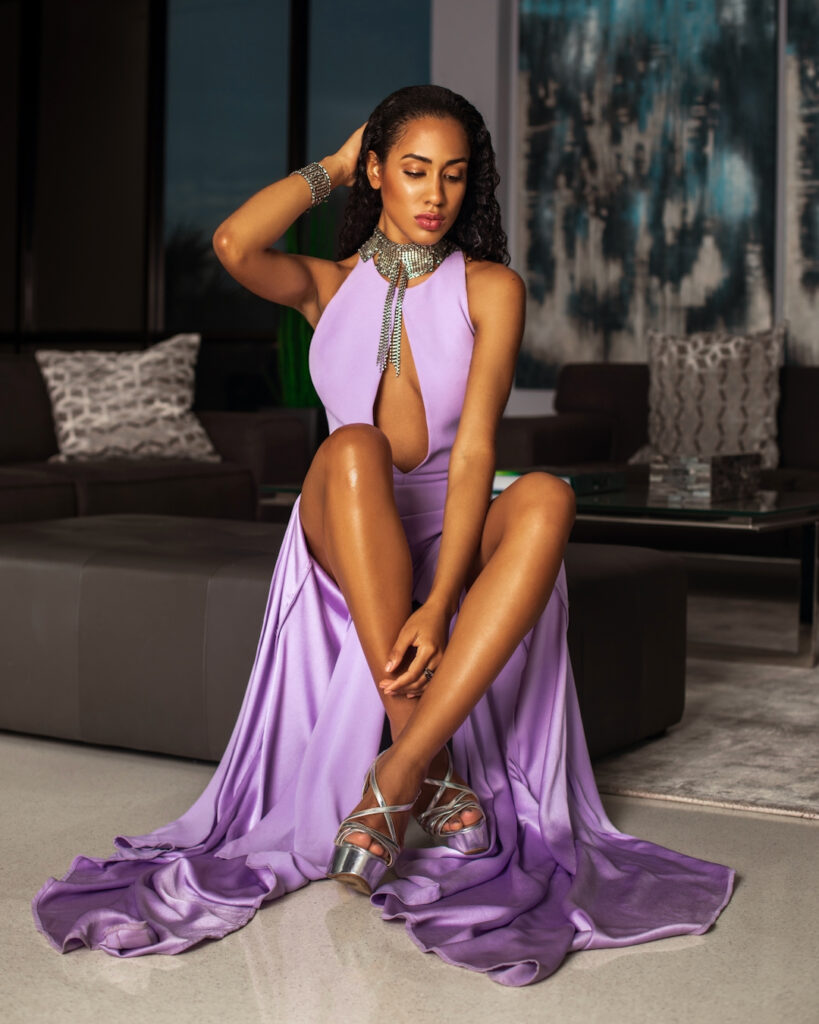 Portrait of a young serene Latin woman with long curly hair and beautiful makeup sitting by herself inside a living room with a window background wearing a purple dress with high heels and jewelry.