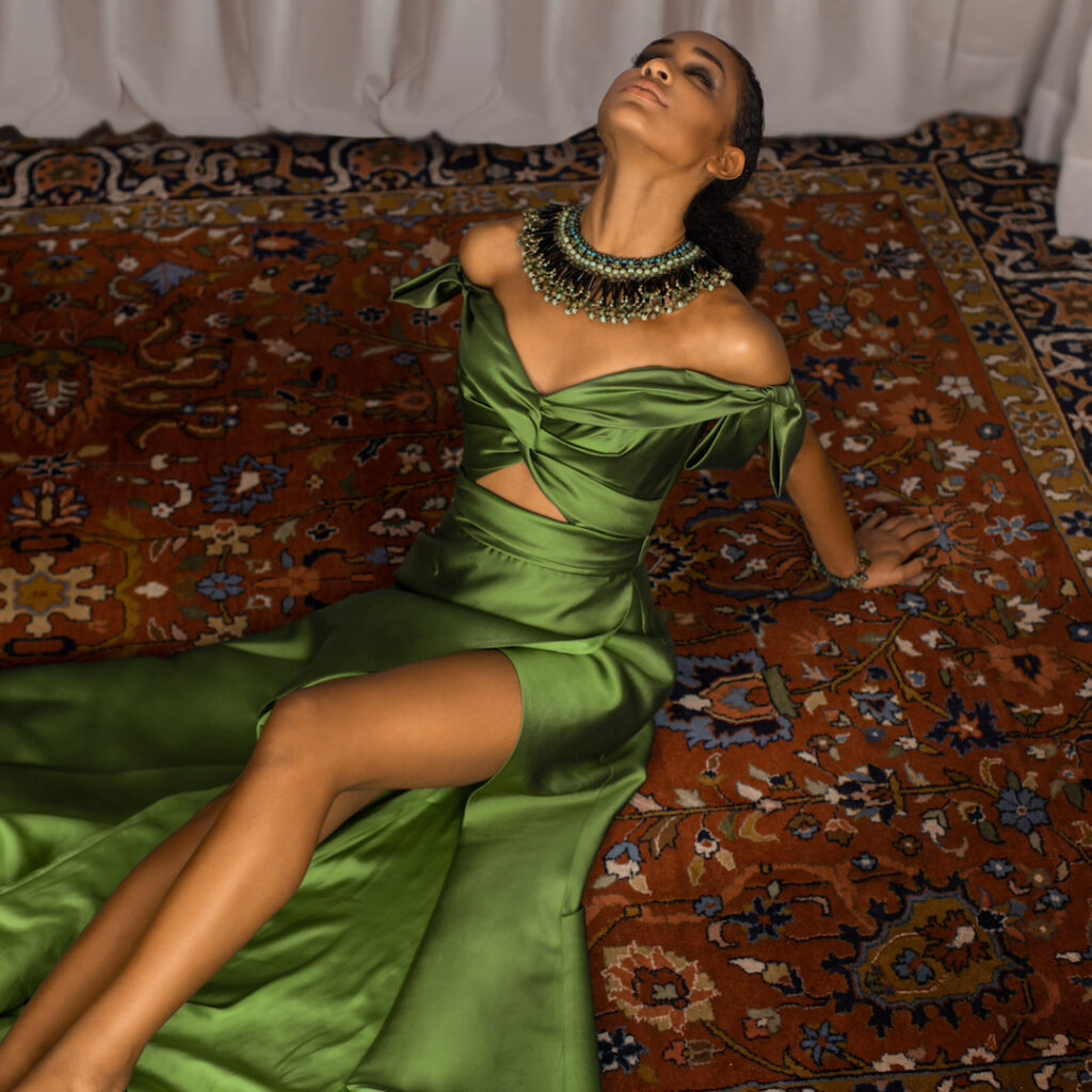 Poise and elegance. Portrait of a young Latin woman with long curly hair and beautiful makeup laying by herself on a Persian rug inside a living room with long white drapes wearing a green dress with jewelry.