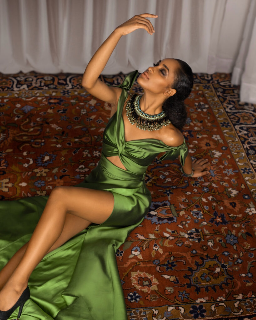 Portrait of an elegant young Latin woman with long curly hair and beautiful makeup laying by herself on a Persian rug inside a living room with long white drapes wearing a green dress with jewelry.