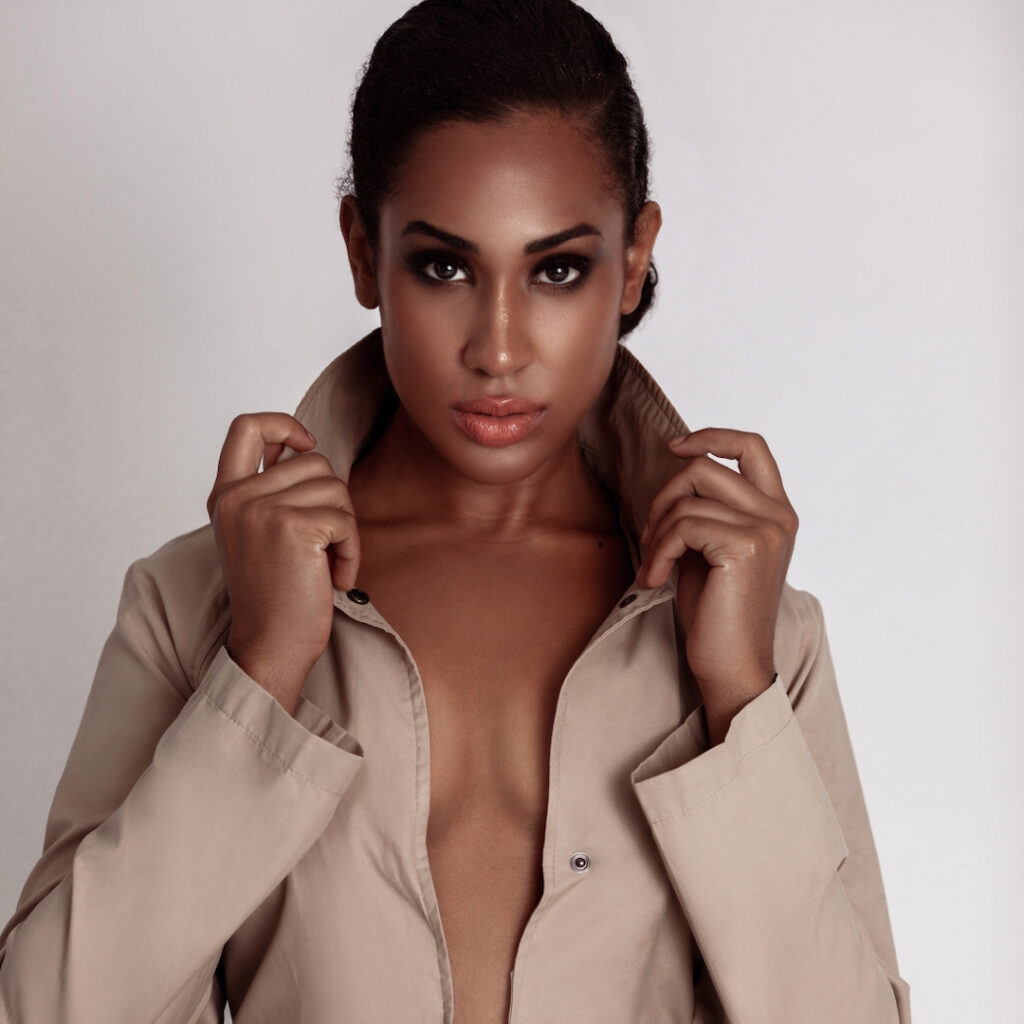 Poise and elegance. Description (200 characters)
Portrait of a young Latin woman with long curly hair and beautiful makeup sitting by herself inside a studio with white background wearing white shorts and a beige trench coat. 