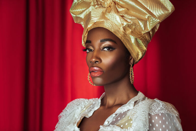 Black designers in fashion. Portrait of a young black female with a gold headpiece & beautiful makeup posing by herself in a studio with red curtain background wearing a white blouse & gold earrings.