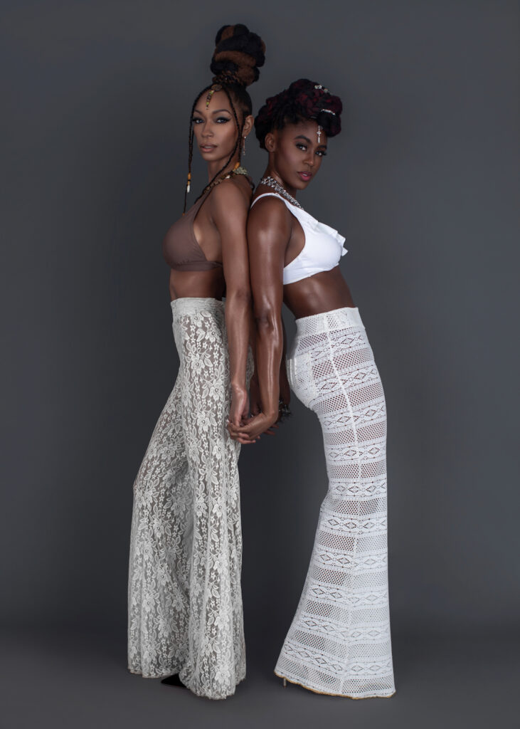 Work Ethic. Portrait of 2 confident Black women with wavy black hair and beautiful makeup jumping by themselves inside a studio with grey background wearing white summer pants with bikini tops and jewelry.
