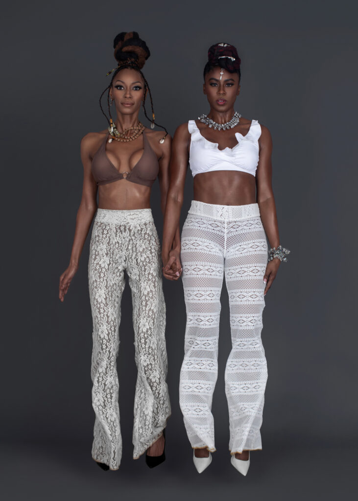Portrait of 2 sensual Black women with wavy black hair and beautiful makeup sitting by themselves inside a studio with grey background wearing white summer pants with bikini tops and jewelry.