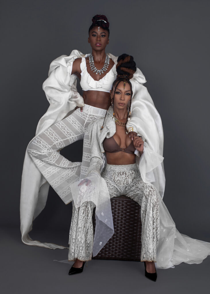 Work Ethic. Portrait of 2 sensual Black women with wavy black hair and beautiful makeup sitting by themselves inside a studio with grey background wearing white summer pants with bikini tops and jewelry.