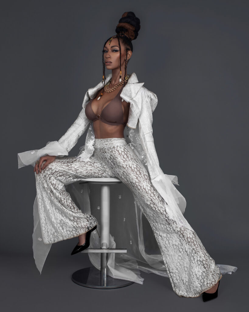 Portrait of a confident Black woman with Updo black hair and beautiful makeup sitting by herself inside a studio with grey background wearing white summer outfit with brown bikini top.