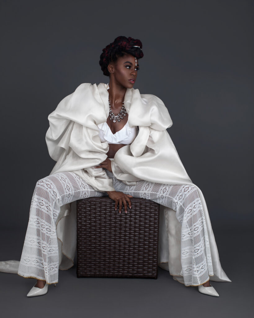 Portrait of an attractive Black woman with Updo black hair and beautiful makeup sitting by herself inside a studio with grey background wearing white summer outfit with bikini top and jewelry.