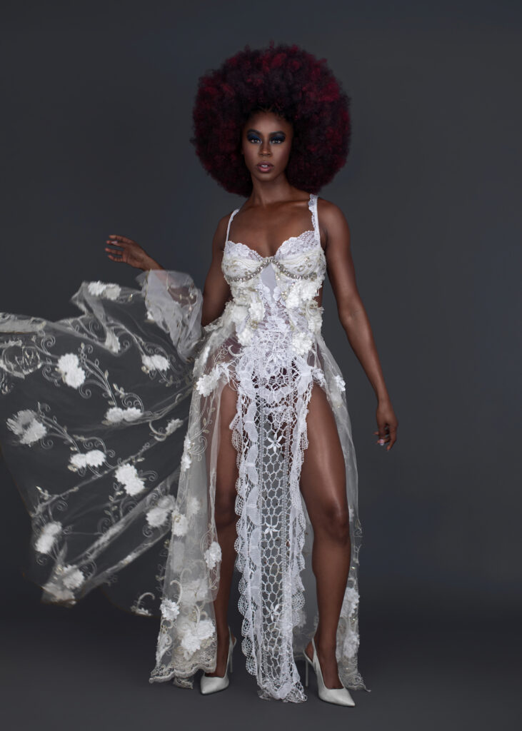 Portrait of a beautiful Black woman with big Afro hair and beautiful makeup standing by herself inside a studio with grey background wearing white summer dress with white high heel shoes.
