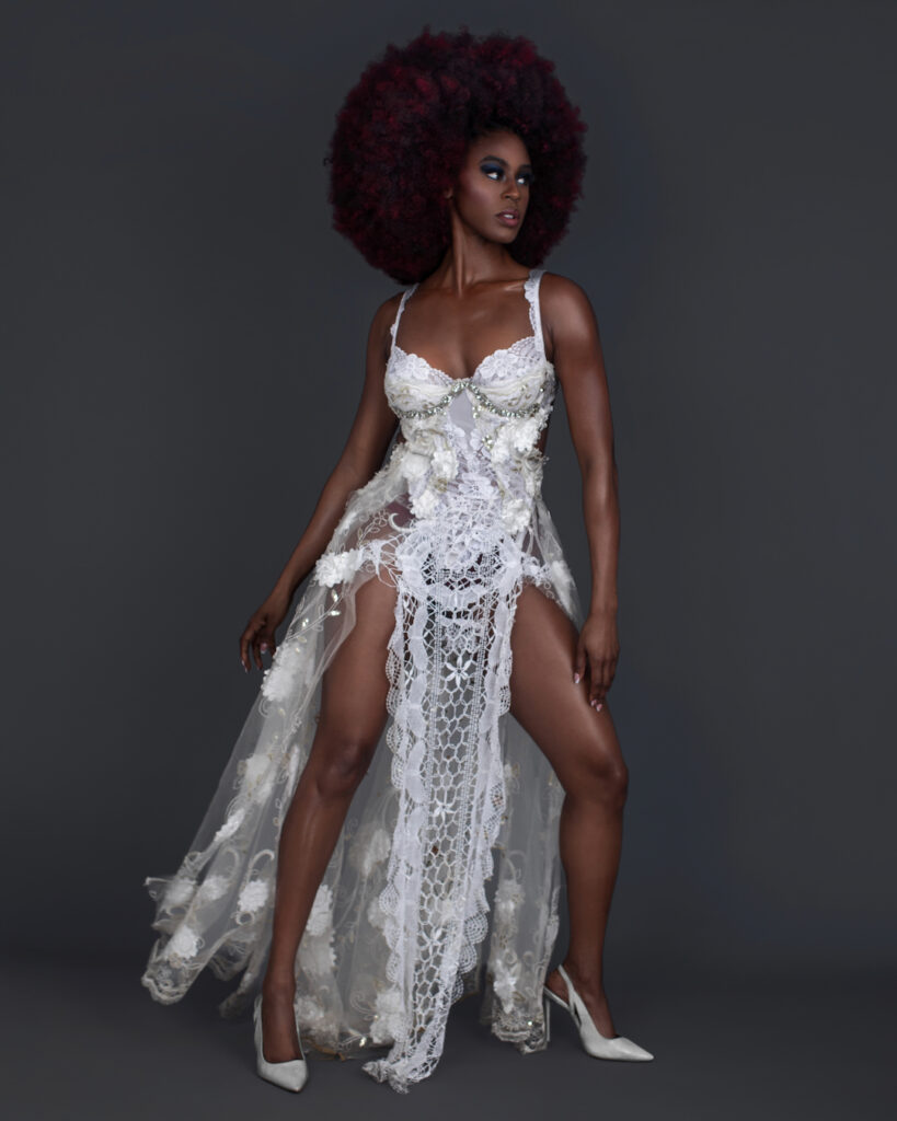 Portrait of a serene Black woman with big Afro hair and beautiful makeup standing by herself inside a studio with grey background wearing white summer dress with white high heel shoes.