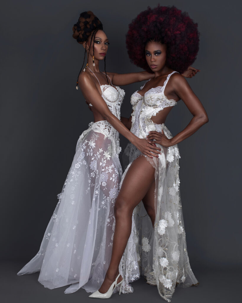 Portrait of 2 confident Black women with wavy black hair and beautiful makeup standing by themselves inside a studio with grey background wearing white summer dresses and jewelry.