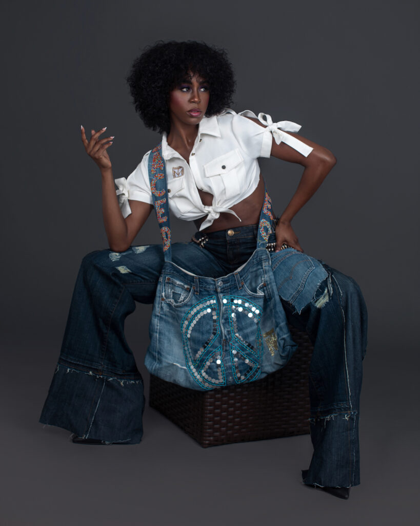 Portrait of a serene Black woman with curly black hair and beautiful makeup sitting by herself inside a studio with grey background wearing denim jeans and bag with white shirt and black high heels.