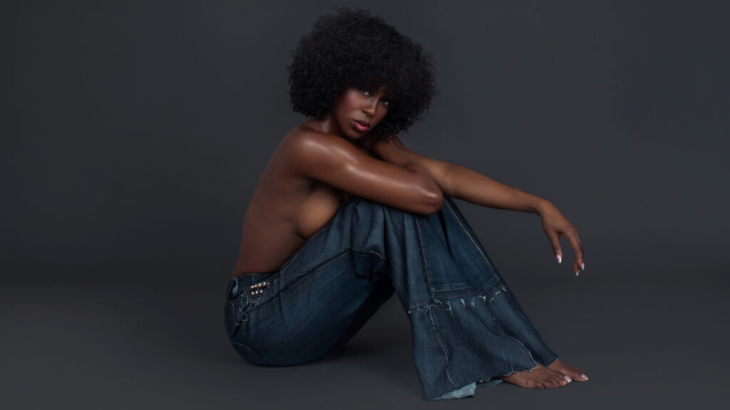 Portrait of a sensual Black woman with curly black hair and beautiful makeup sitting by herself inside a studio with grey background wearing denim jeans.