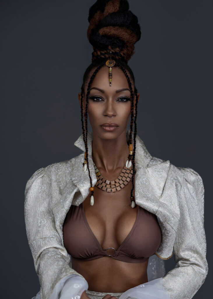 Work Ethic. Portrait of a beautiful Black woman with Updo black hair and beautiful makeup sitting by herself inside a studio with grey background wearing white jacket with brown bikini top and jewelry.
