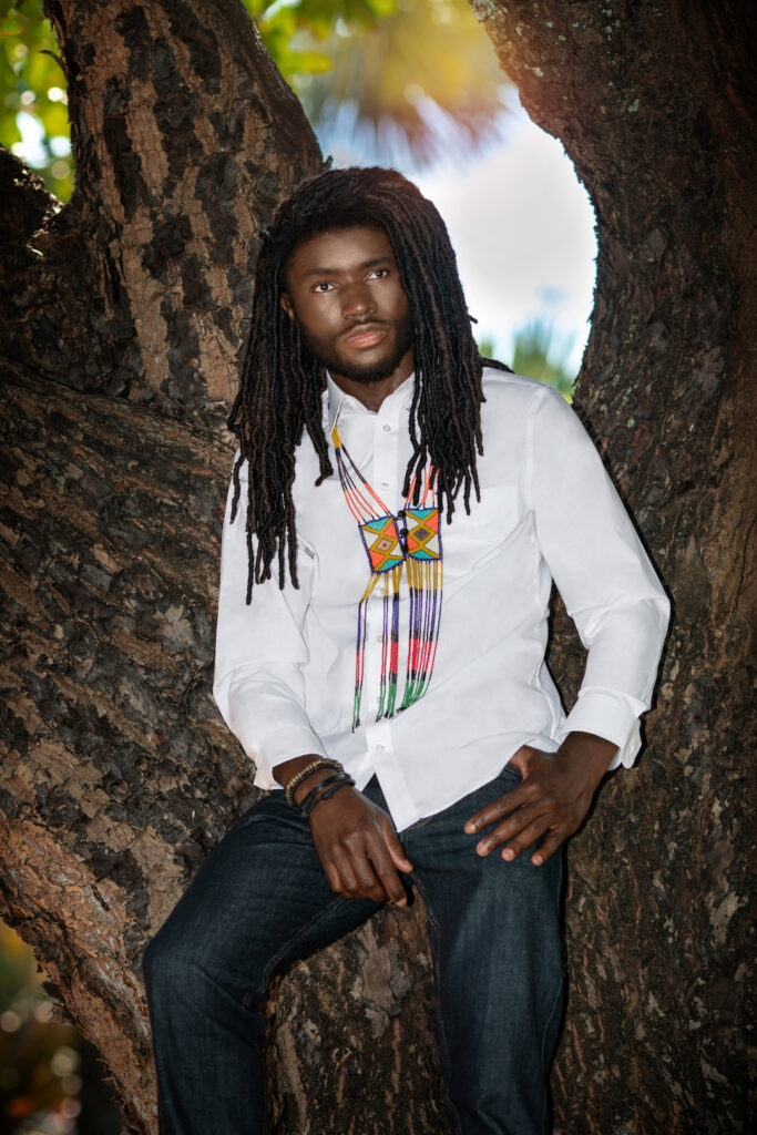 Closeup portrait of a serene Millennials black man with long dreadlocks posing by himself outside leaning on a very old oak tree wearing blue jeans, white long sleeve shirt and African jewelry.